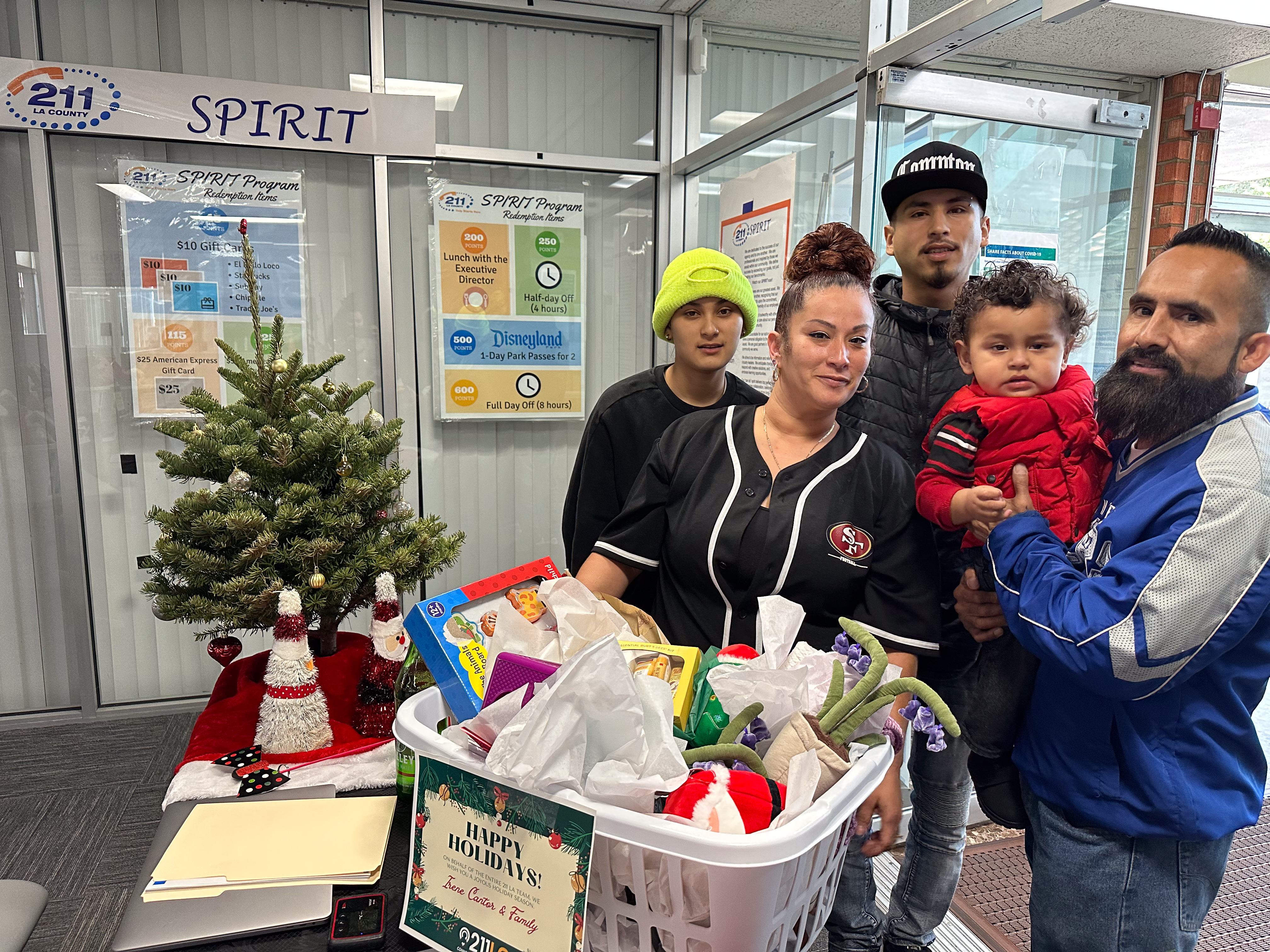 A family is standing next to a Christmas tree.  The family is holding a large hamper filled with what appears to be gifts or toys. A sign in the basket says "HAPPY HOLIDAYS" The man on the right is wearing a blue and white jacket and holding a baby wearing a red vest. The woman in the center is wearing a black and white shirt with the "49ers" logo on it.