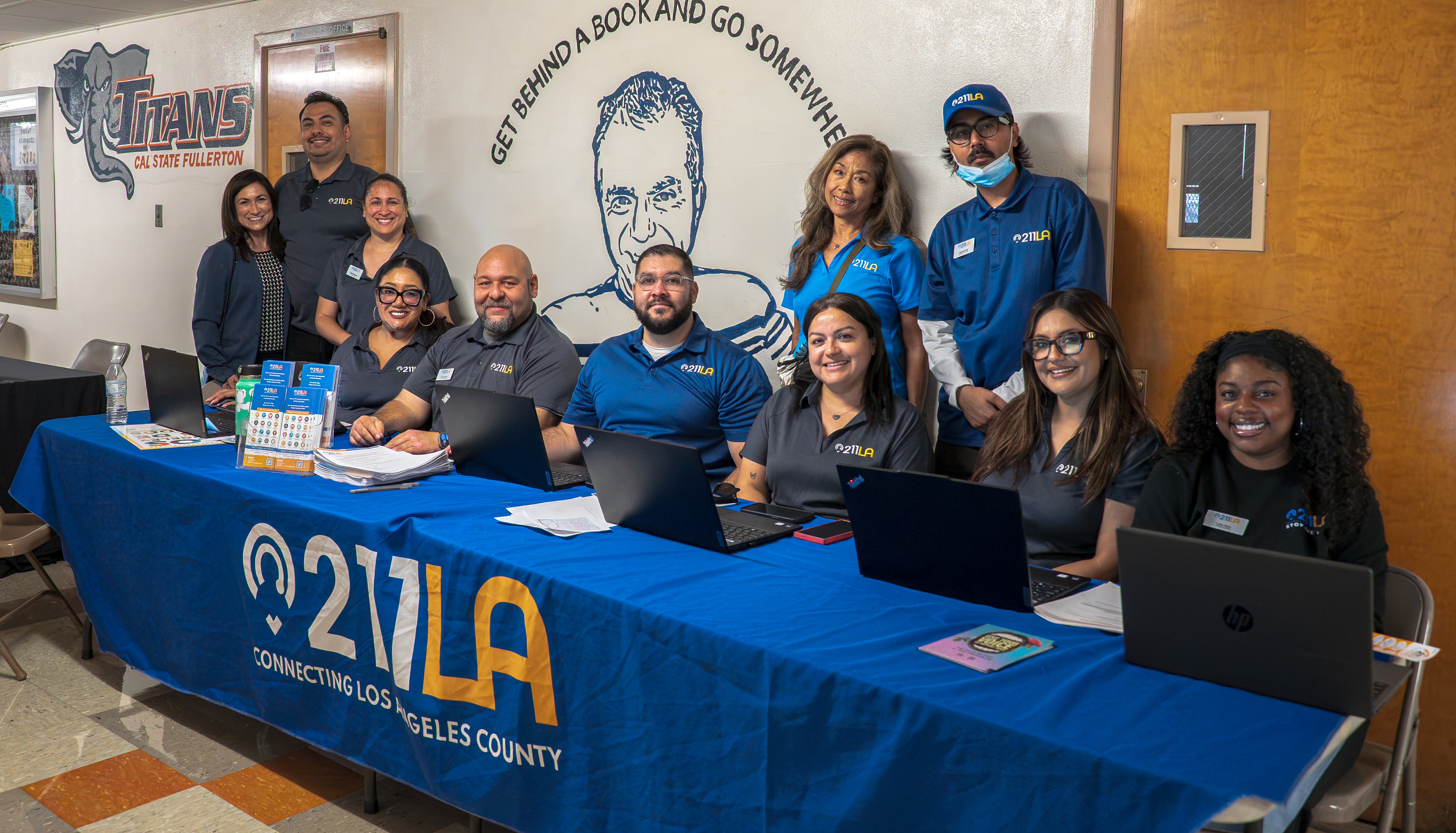 Ten 211 LA staff members wearing matching branded shirts smile behind an event resource table equipped with laptops and flyers, set against a wall with school murals.