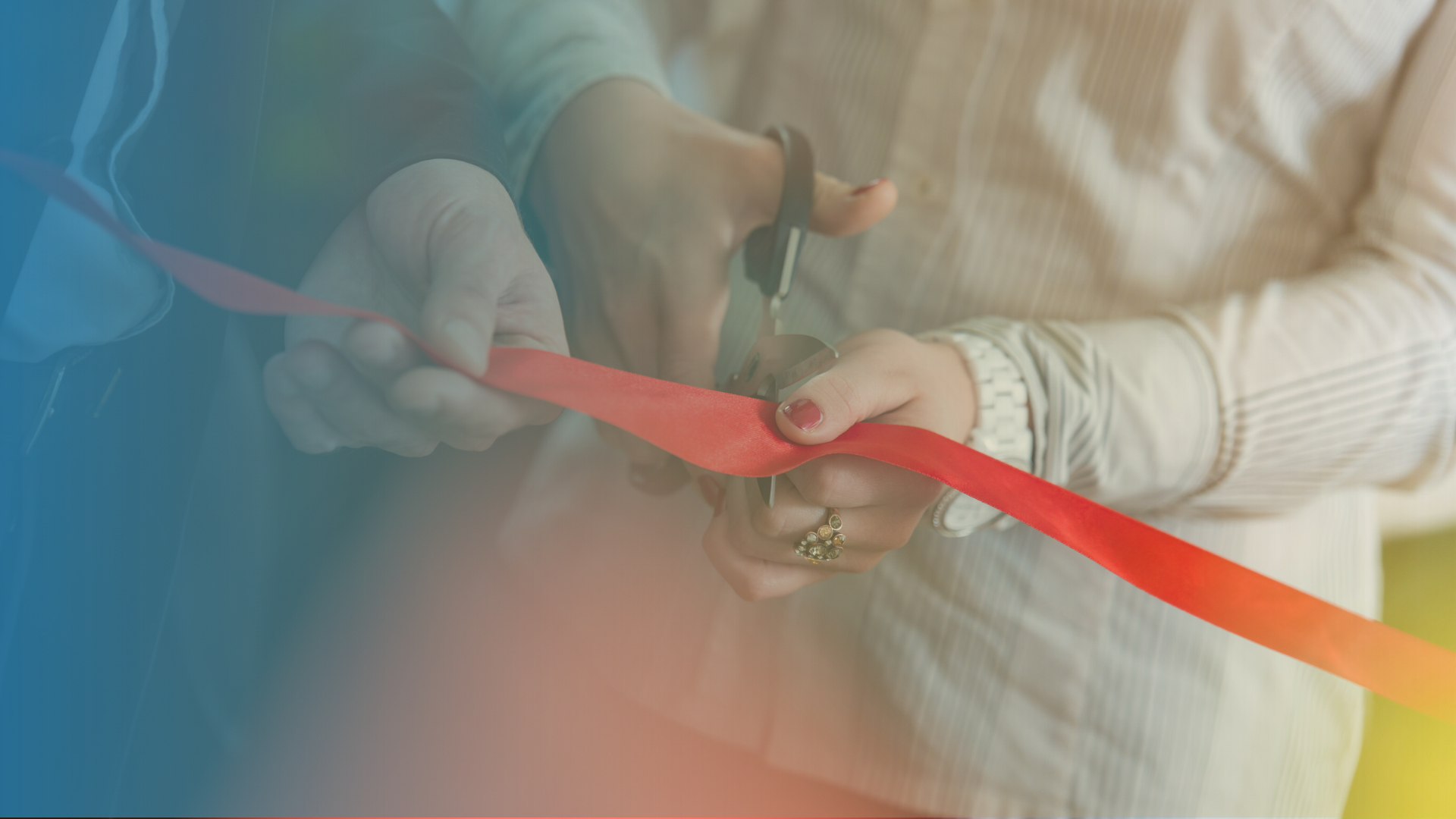 Close-up of two people's hands participating in a ribbon-cutting ceremony, with one person holding a red ribbon taut while the other cuts it with a pair of scissors.