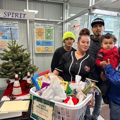 A family is standing next to a Christmas tree.  The family is holding a large hamper filled with what appears to be gifts or toys. A sign in the basket says "HAPPY HOLIDAYS" The man on the right is wearing a blue and white jacket and holding a baby wearing a red vest. The woman in the center is wearing a black and white shirt with the "49ers" logo on it.