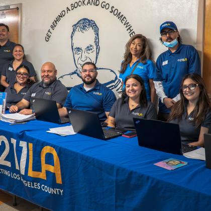 Ten 211 LA staff members wearing matching branded shirts smile behind an event resource table equipped with laptops and flyers, set against a wall with school murals.