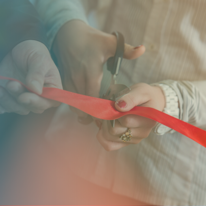Close-up of two people's hands participating in a ribbon-cutting ceremony, with one person holding a red ribbon taut while the other cuts it with a pair of scissors.