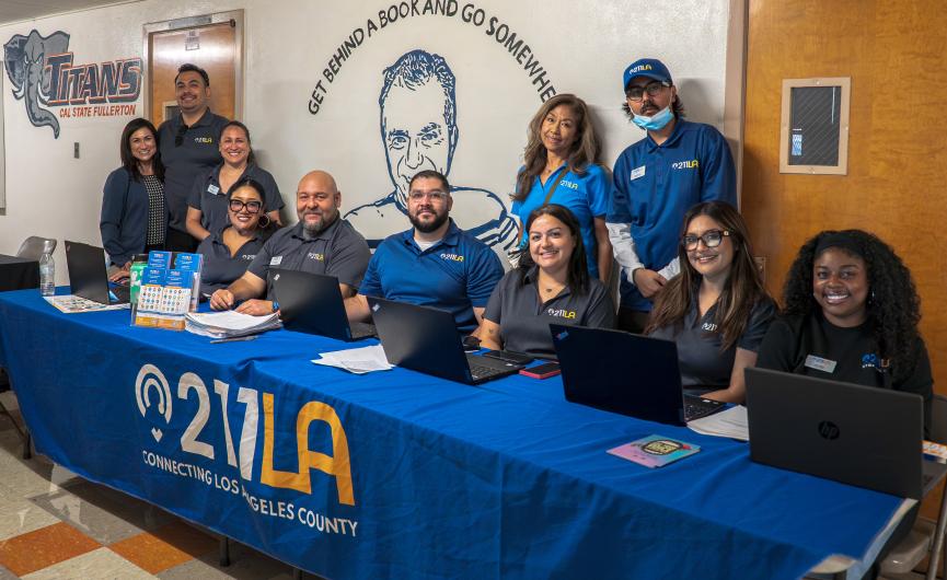 Stevenson Middle School, ten 211 LA staff members wearing matching branded shirts smile behind an event resource table equipped with laptops and flyers, set against a wall with school murals.