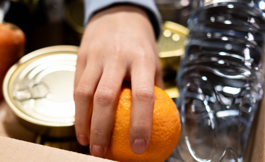 Person reaching into a box of food such as canned food, water bottle, and orange. Person is directly reaching for orange.