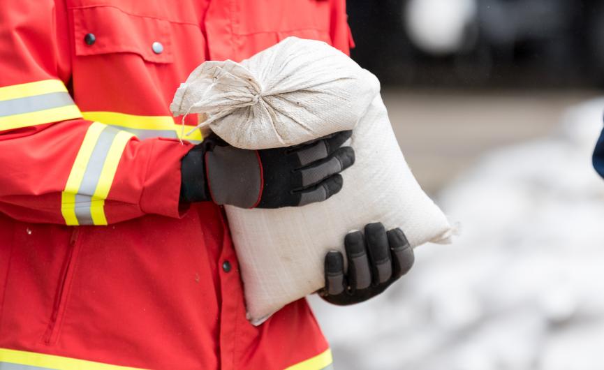 A person wearing a red high-visibility jacket and black gloves holding a white sandbag.