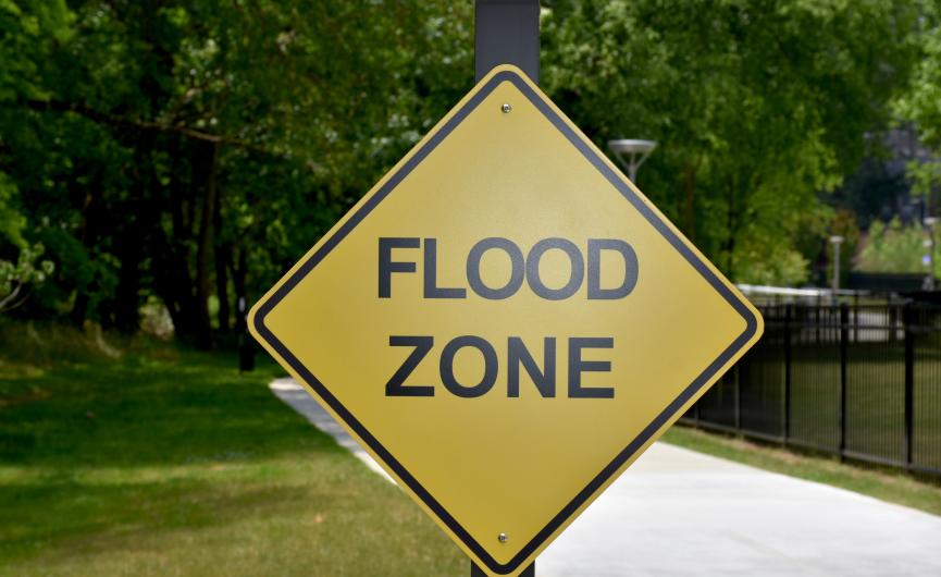 A yellow diamond-shaped warning sign that reads "FLOOD ZONE" next to a paved path.