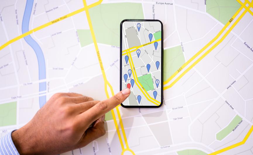 A hand pointing at a red location pin on a smartphone map, with the phone resting on top of a printed paper map.
