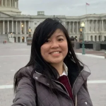 Becky Cheng Smiling wearing a grey puffer jacket with long black hair standing outside with structures around 