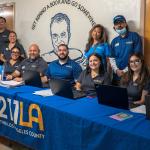 Ten 211 LA staff members wearing matching branded shirts smile behind an event resource table equipped with laptops and flyers, set against a wall with school murals.